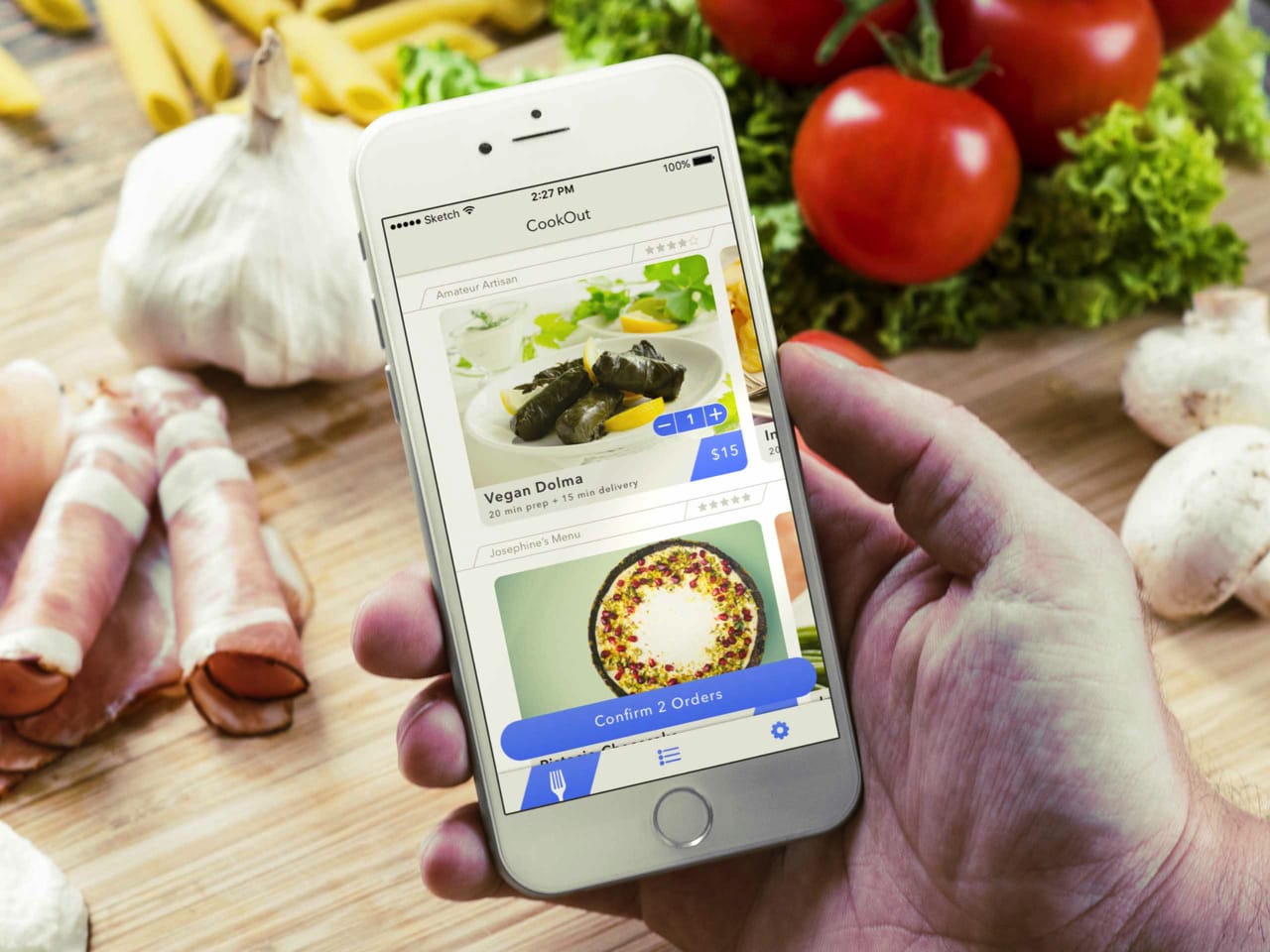 a person holding an iphone with the cookout app showing on the screen. the background of the photo is a kitchen table top with food ingredients on it . 